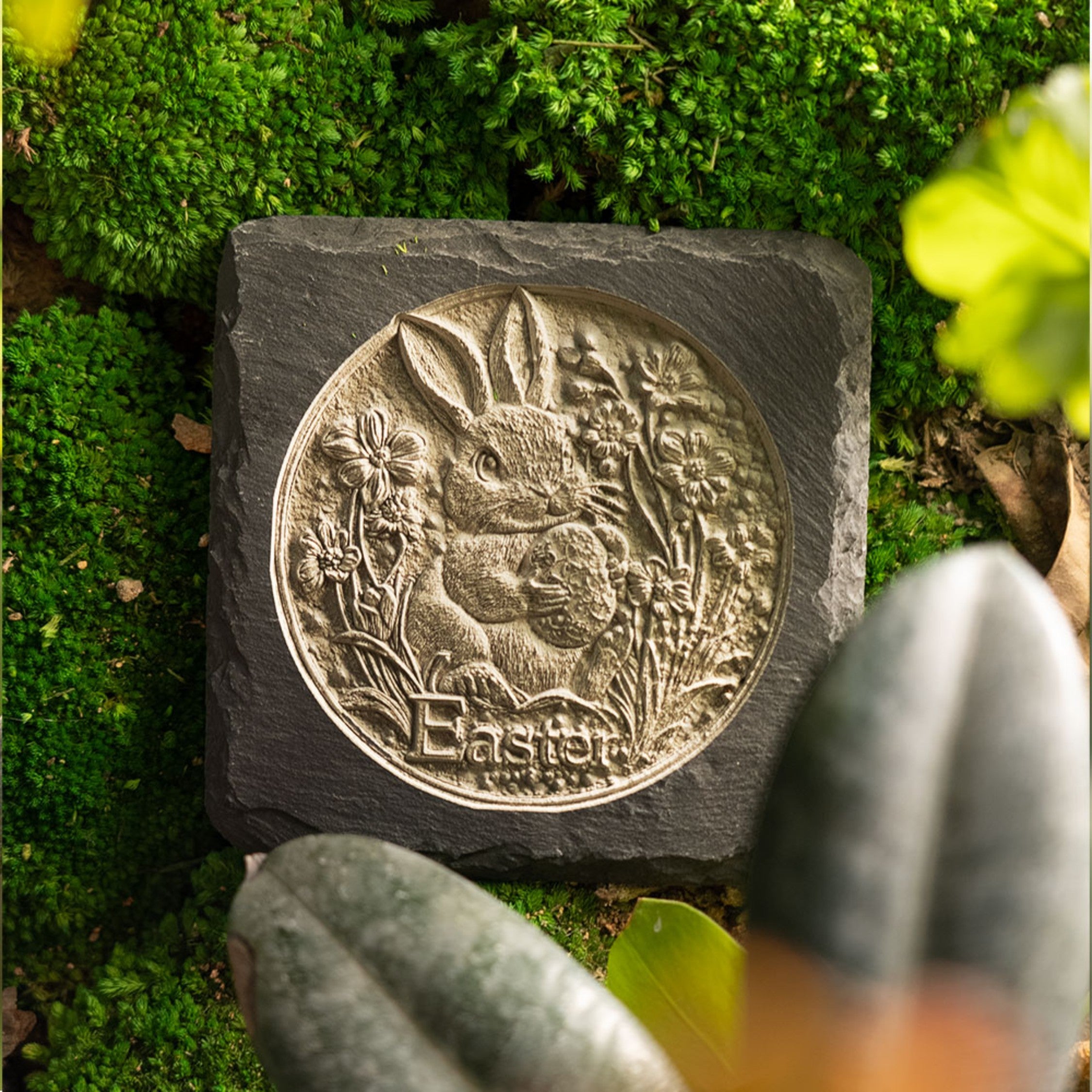 A laser-engraved slate plaque featuring a highly detailed, 3D-style circular Easter design of a rabbit holding an egg, surrounded by spring flowers and foliage, with the word "Easter" engraved at the bottom. It’s presented as a durable, decorative piece perfect for seasonal home or garden decor.