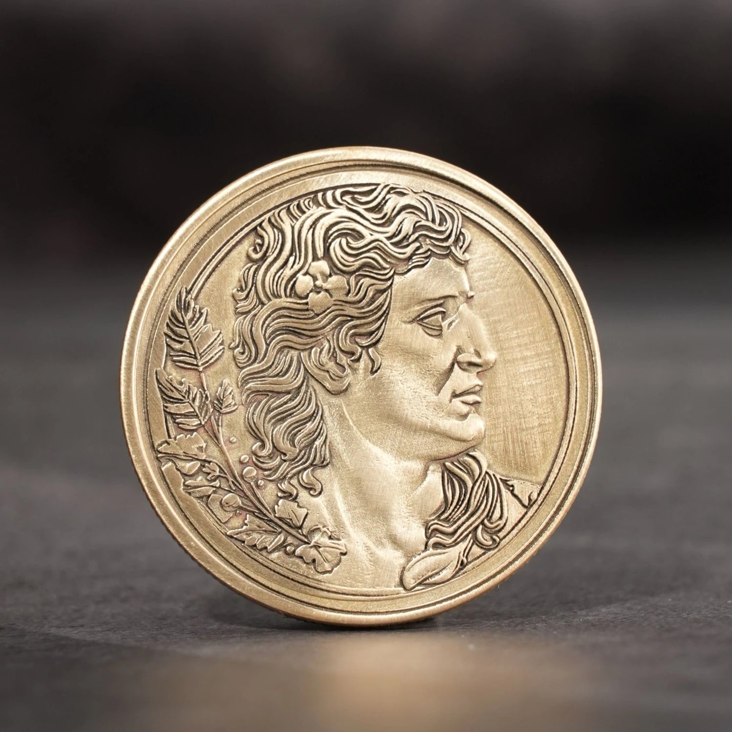 A laser-engraved gold metal coin featuring a highly detailed profile of a classical figure with laurel wreath accents, showcasing the intricate precision achievable with laser engraving on blank metal coin blanks, presented against a dark, textured backdrop.