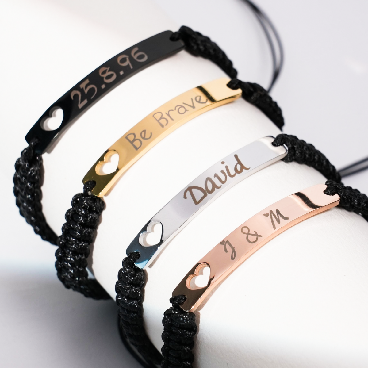 A stack of four adjustable personalized bracelets with black braided cords, featuring metal plates in black, gold, silver, and rose gold finishes. Each plate is custom-engraved with unique details (a date "25.8.96", the phrase "Be Brave", the name "David", and initials "J & M") and accented with a small heart cutout.