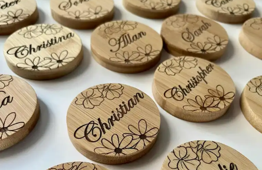 Real User Stories: Assortment of custom laser-engraved wooden circular pieces (with names like Christina, Allan and floral patterns) from Mia's Etsy Store; Mia shares the LaserPecker LP5 is a business gamechanger—easy to bring to vendor events, equipped with camera enclosure & auto alignment, and offers endless material possibilities.