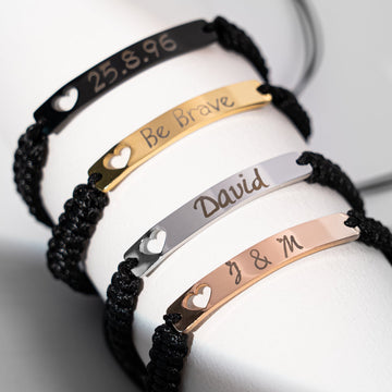A set of four personalized laser-engraved stainless steel bar bracelets with black braided cords, featuring custom engravings including the date "25.8.96", the phrase "Be Brave", the name "David", and initials "J & M", demonstrating the versatility of these bracelet blanks for creating meaningful, custom laser-engraved accessories.