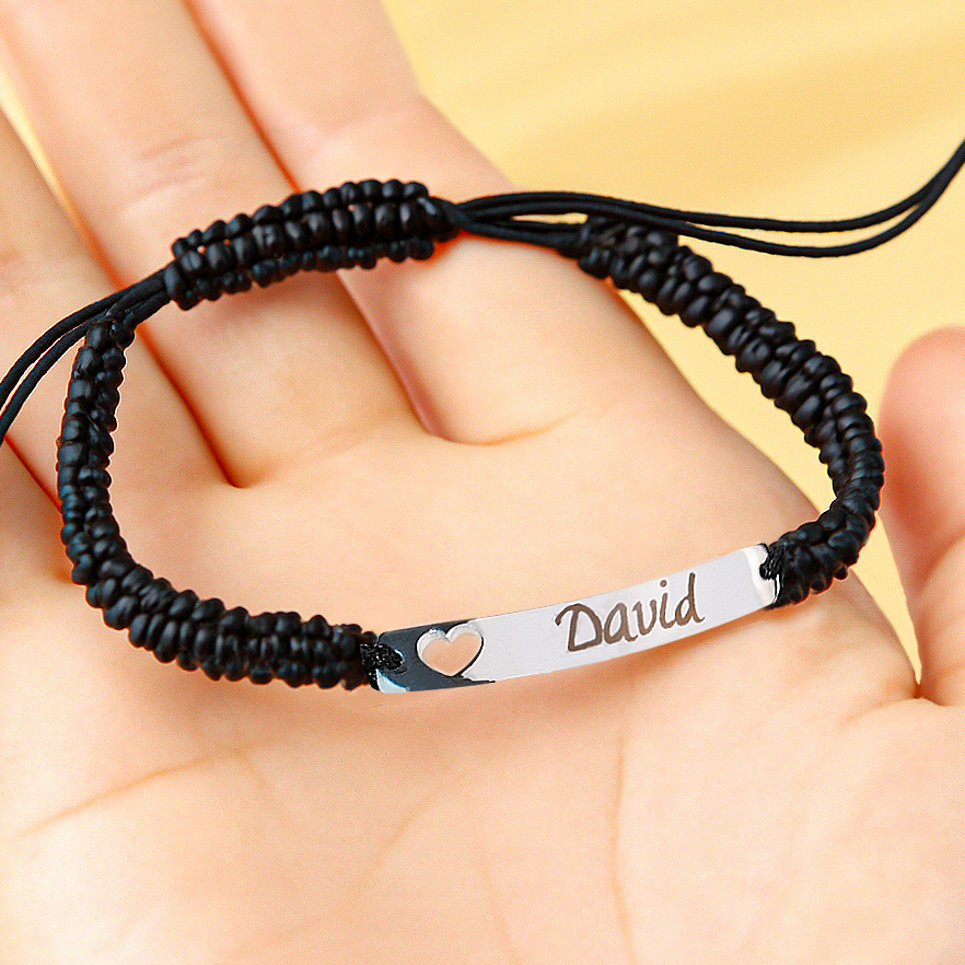 A personalized adjustable bracelet displayed on an open palm, featuring a black braided cord and a polished stainless steel plate engraved with the name "David", accented by a small heart cutout on the metal tag.