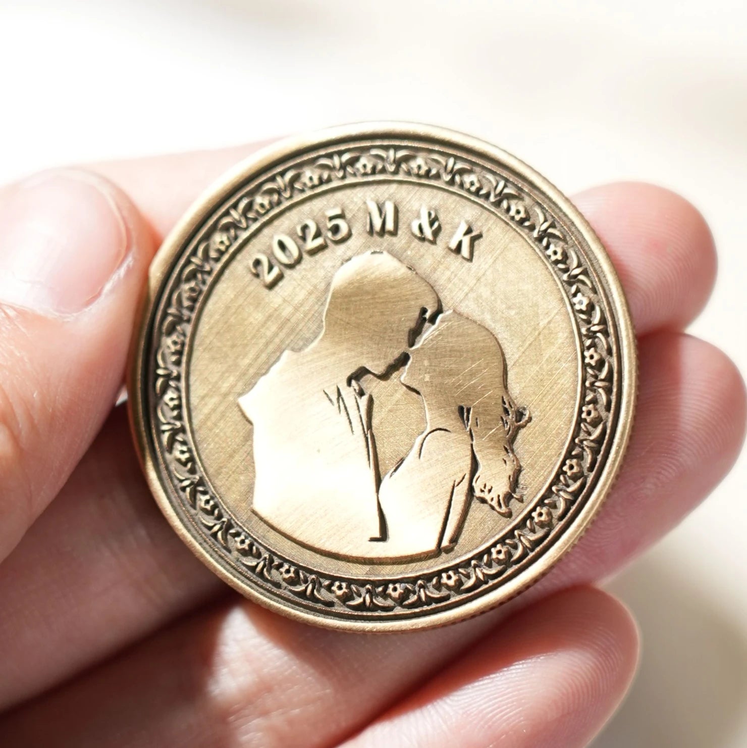 A hand-held, custom laser-engraved bronze coin featuring a silhouette of a kissing couple, the year "2025", and the initials "M & K", showcasing a personalized keepsake created from a blank metal coin.