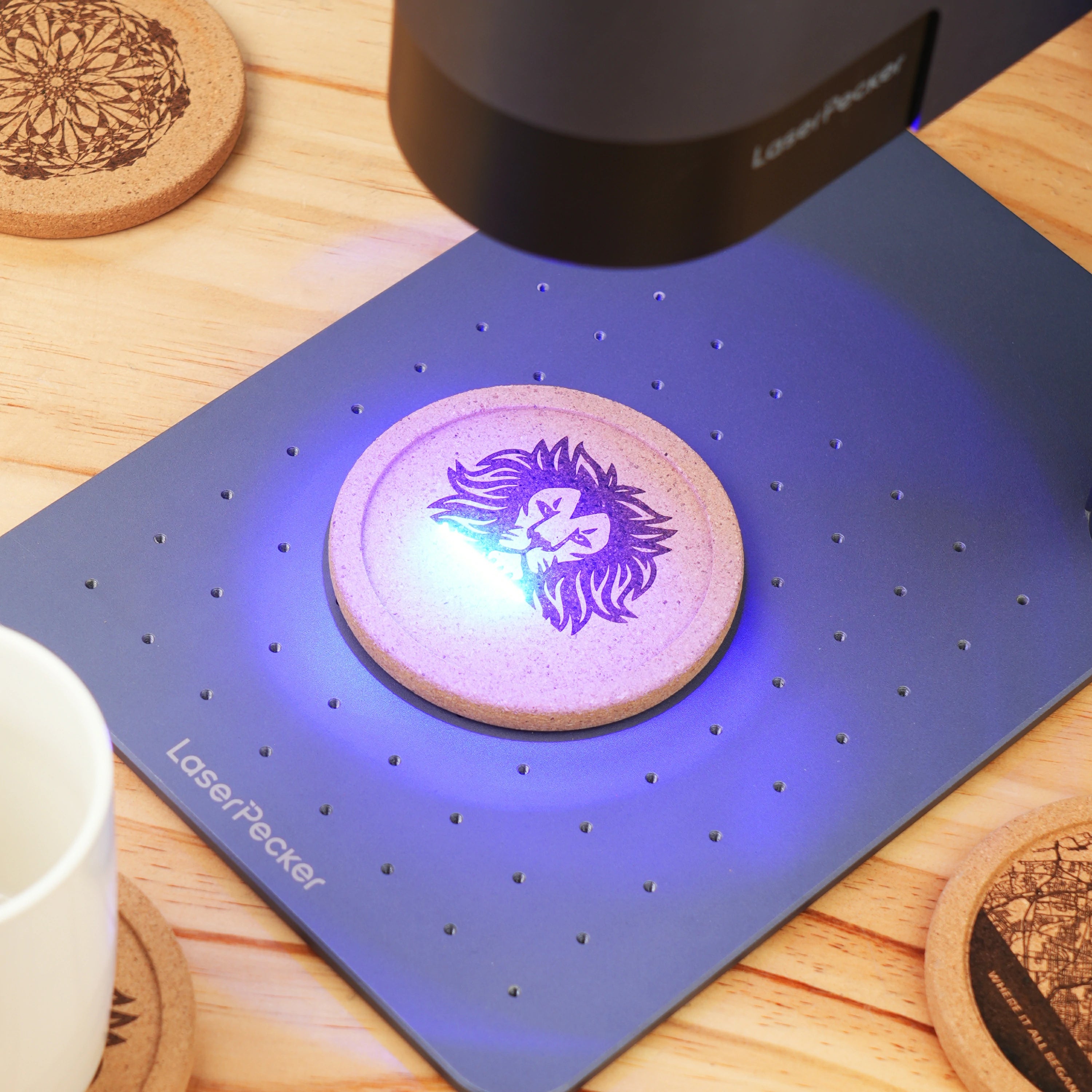 A LaserPecker laser engraver in action, engraving a detailed lion head design onto a round cork coaster. The setup includes a blue engraving mat, with finished coasters featuring mandala and cityscape designs visible in the background, demonstrating the use of laser engraving to create personalized home accessories.
