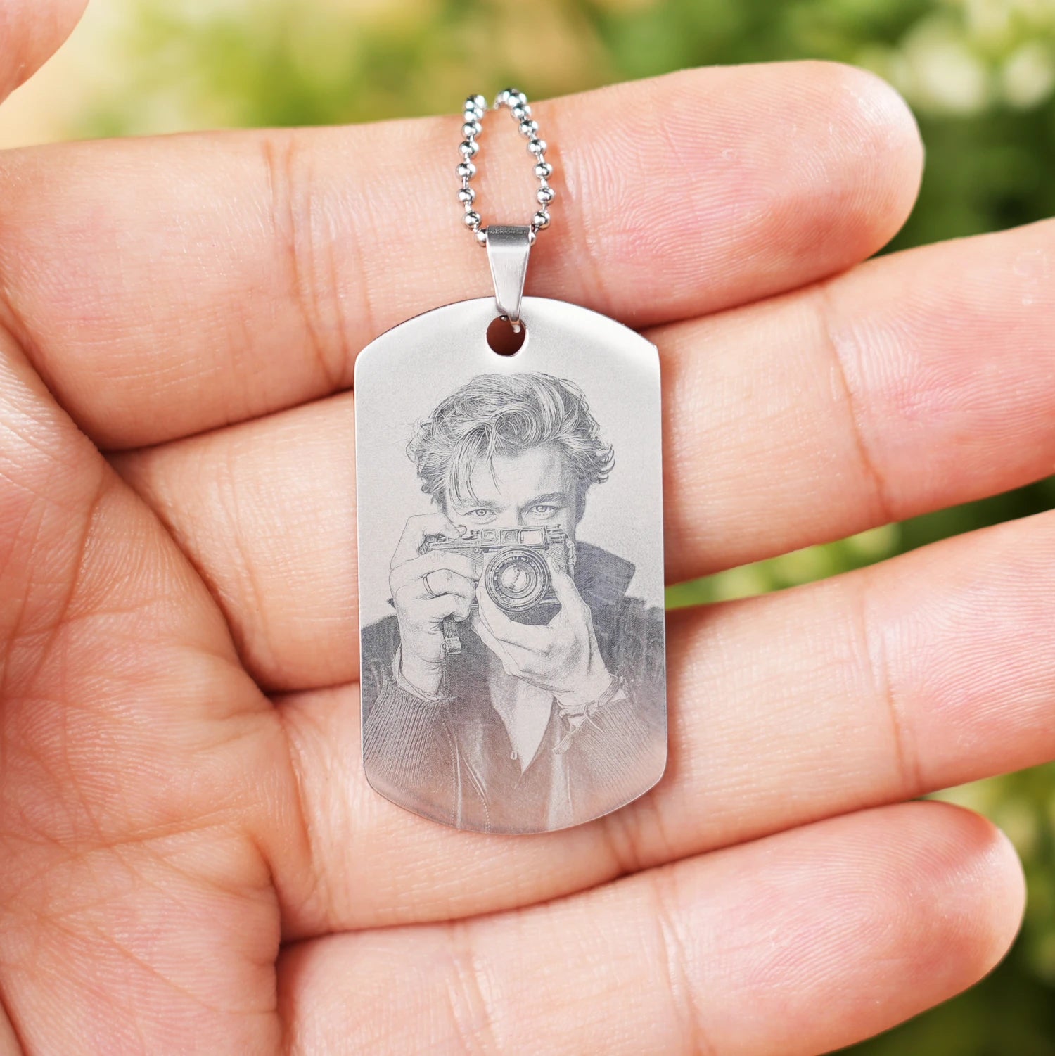 A personalized stainless steel dog tag necklace held in an open palm, featuring a high-detail laser-engraved portrait of a person holding a camera, paired with a classic beaded ball chain.