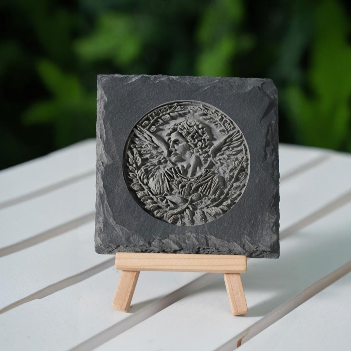 A laser-engraved slate plaque featuring a highly detailed, 3D-style circular portrait of a classical figure surrounded by foliage, displayed on a small wooden easel. The natural, rough edges of the slate contrast with the intricate, sculptural details of the engraving, creating an elegant, durable decorative piece with an artistic, timeless feel.
