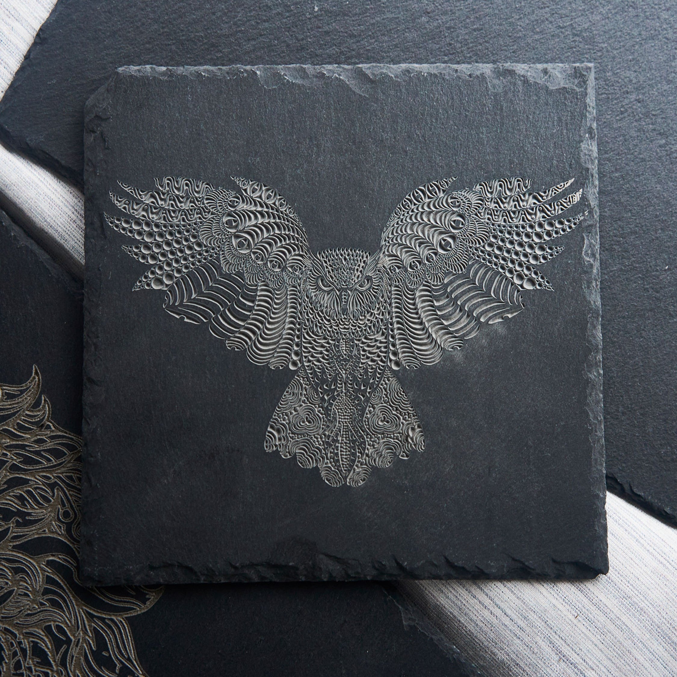 A laser-engraved slate coaster featuring an intricate, abstract eagle design composed of swirling, textured patterns that create a sense of depth and movement. The high-contrast engraving stands out against the dark slate, making it a striking decorative piece or functional drinkware accessory for modern or artistic home decor.