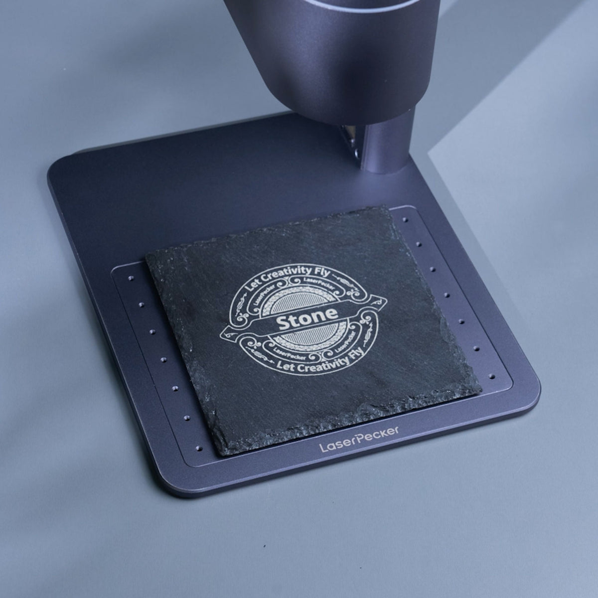 A LaserPecker laser engraver engraving a decorative vintage-style logo onto a square slate coaster. The design features the word "Stone" at its center, surrounded by the slogan "Let Creativity Fly" and intricate scrollwork, showcasing the machine’s precision for creating detailed, professional-looking branding on natural stone surfaces.