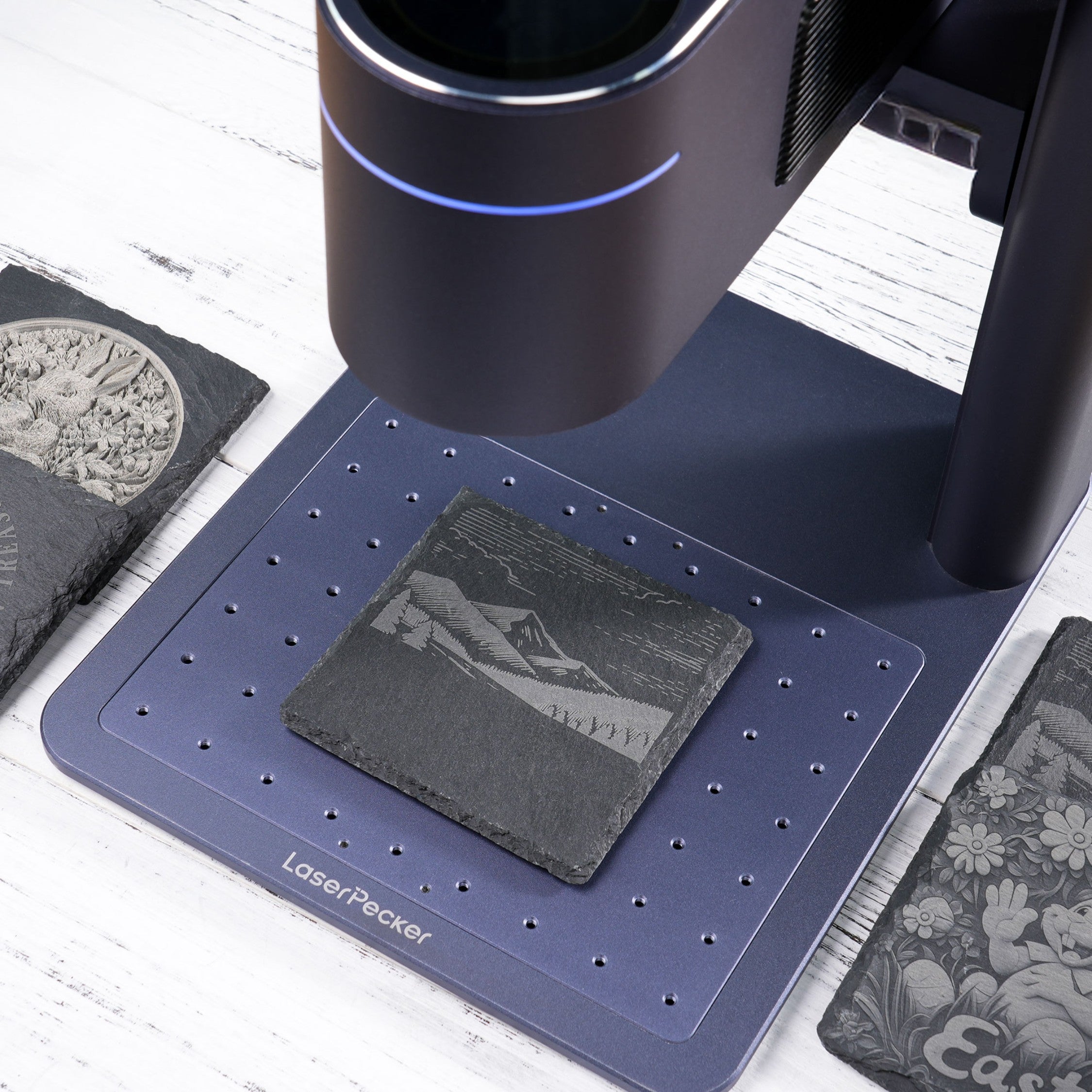 A LaserPecker laser engraver in action, engraving a detailed mountain landscape onto a square slate coaster. The setup is on a branded blue mat, with finished slate pieces featuring intricate designs visible nearby, demonstrating the machine’s precision for creating personalized slate decor and accessories.