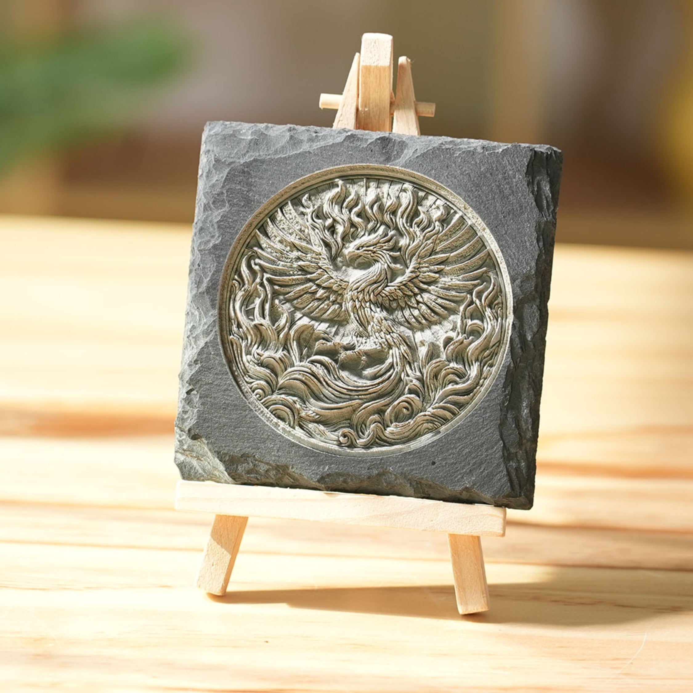 A laser-engraved slate plaque featuring a highly detailed, 3D-style phoenix design, displayed on a small wooden easel. The natural rough edges of the slate contrast with the intricate, flame-like details of the phoenix, creating an elegant and durable decorative piece that symbolizes rebirth and strength.
