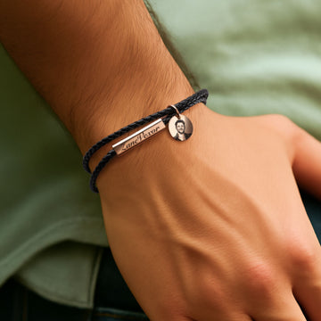 A personalized double-layered braided leather bracelet worn on a man’s wrist, featuring a rectangular metal bar engraved with the name "Lane Dexor" and a small circular charm with a laser-engraved portrait, designed as a custom sentimental gift.