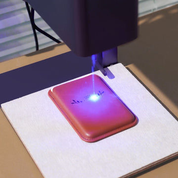 This image captures a laser engraving machine in action, its blue laser beam actively etching a delicate design onto a brown faux leather card holder. The precision of the laser creates crisp, permanent details, showcasing how functional accessories can be transformed into personalized gifts or branded items with custom text, logos, or patterns.

