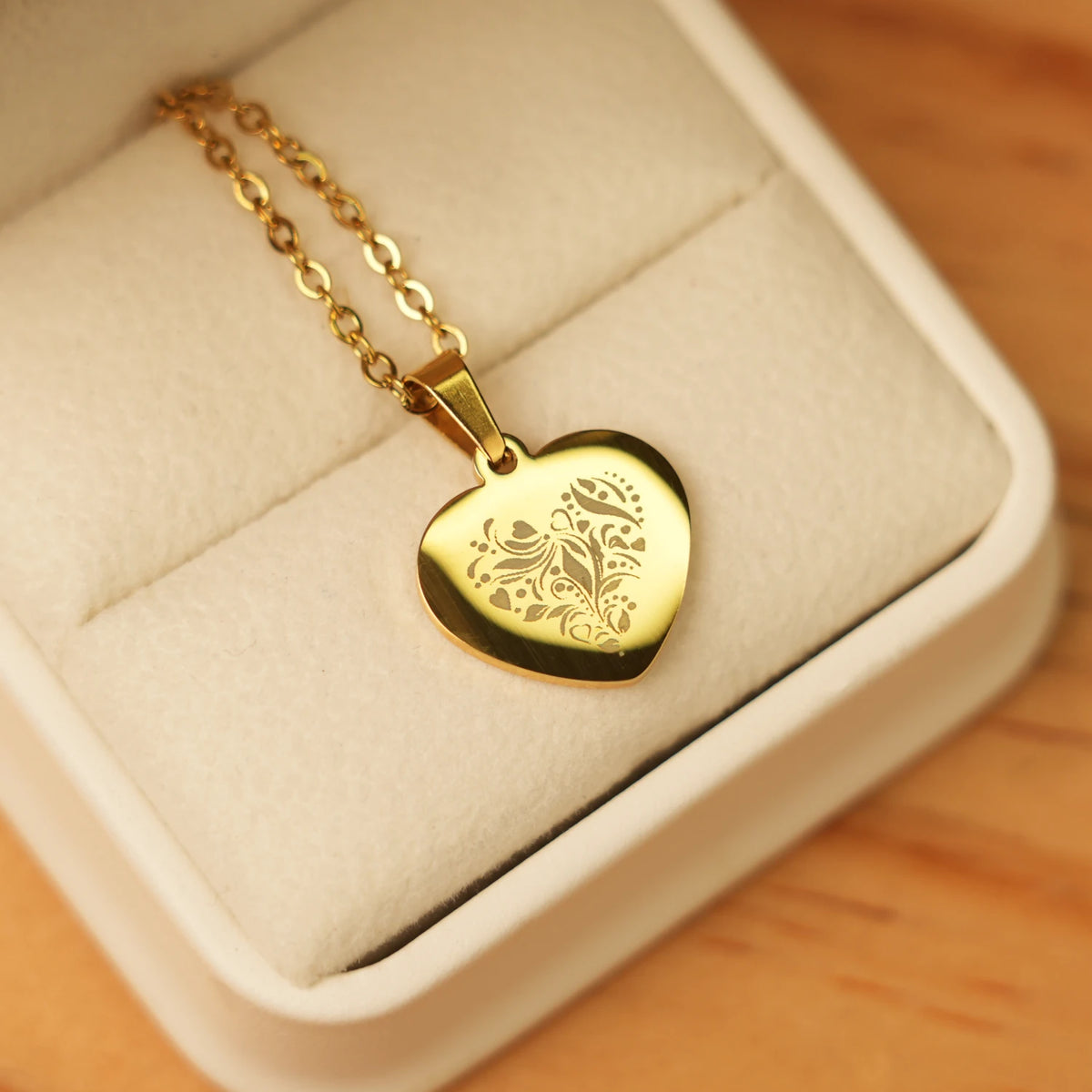 This elegant gold heart-shaped pendant necklace rests in a soft, cream-colored jewelry box, showcasing a delicate, laser-engraved floral and scrollwork pattern that adds a touch of timeless sophistication. The polished, reflective surface of the heart pendant contrasts beautifully with its intricate, matte engraving, making it a refined gift for occasions like birthdays, anniversaries, or Valentine’s Day, where personalization and classic style are key.
