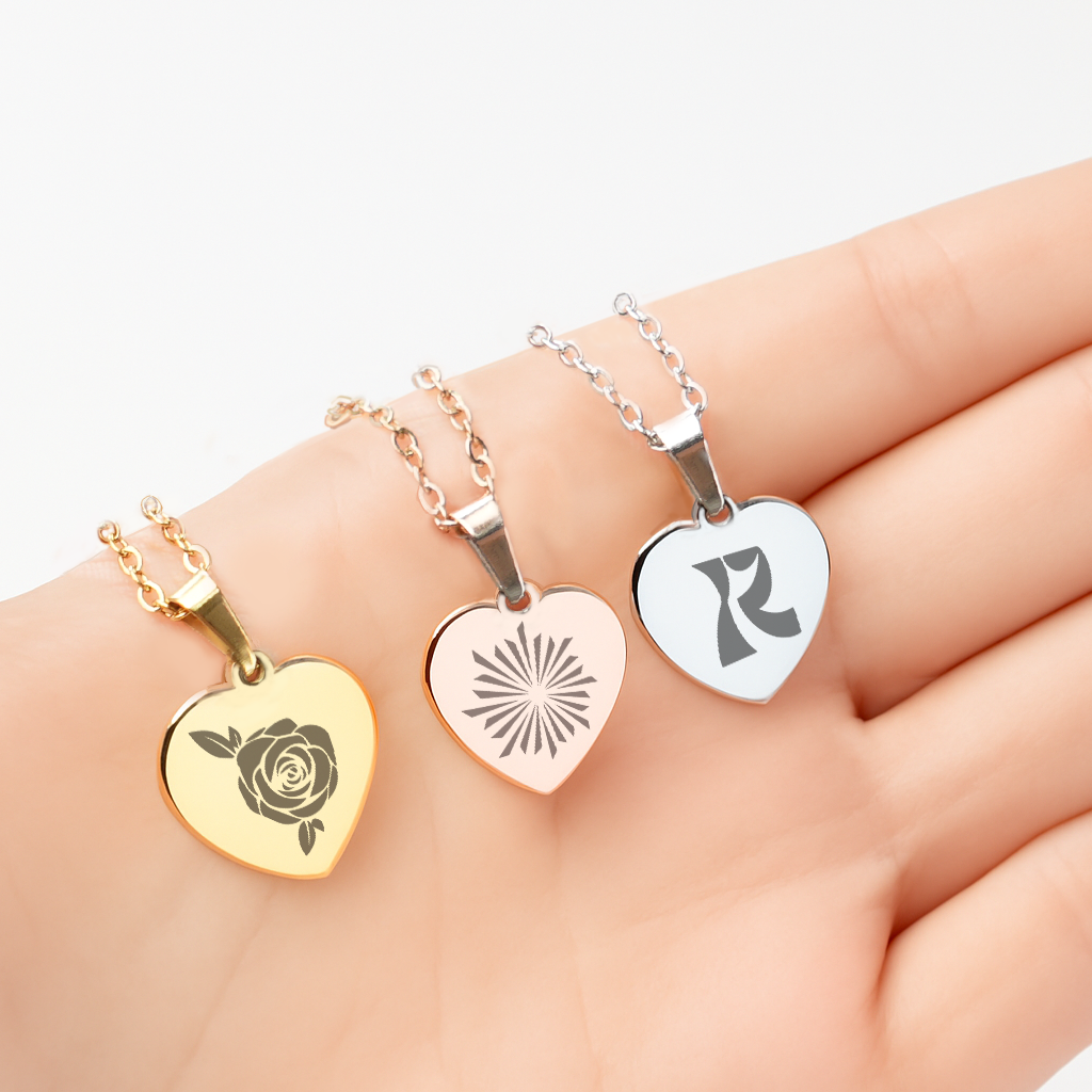 Three delicate heart-shaped pendant necklaces, each finished in a distinct polished metal and personalized with a unique laser-engraved design: a gold necklace features a classic rose pattern, a rose gold version displays a modern sunburst motif, and a silver pendant bears a stylized initial "R". These customizable pieces exemplify how blank metal jewelry can be transformed into meaningful, one-of-a-kind gifts for special occasions like anniversaries, birthdays, or Valentine’s Day.